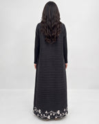 A woman viewed from behind wearing a long black ribbed dress with long sleeves and a white floral embroidered border at the hem, against a plain light gray studio background.