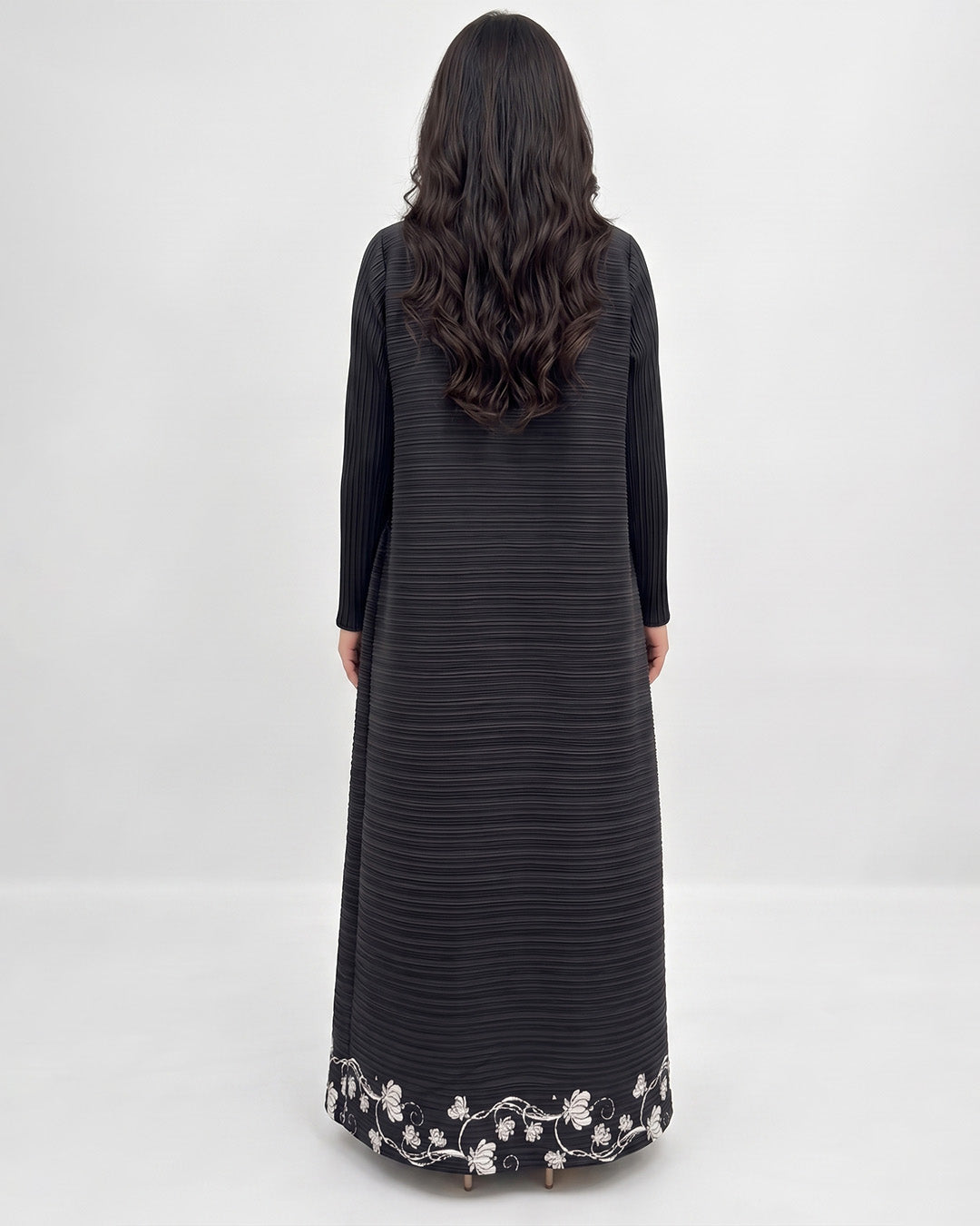 A woman viewed from behind wearing a long black ribbed dress with long sleeves and a white floral embroidered border at the hem, against a plain light gray studio background.
