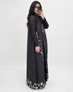 A woman takes a mirror selfie wearing a long black pleated abaya dress with white floral embroidery along the edges and hem, set against a plain light gray studio background.