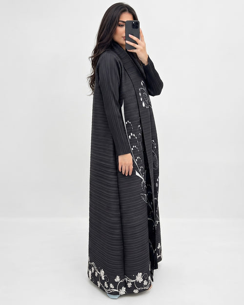 A woman takes a mirror selfie wearing a long black pleated abaya dress with white floral embroidery along the edges and hem, set against a plain light gray studio background.