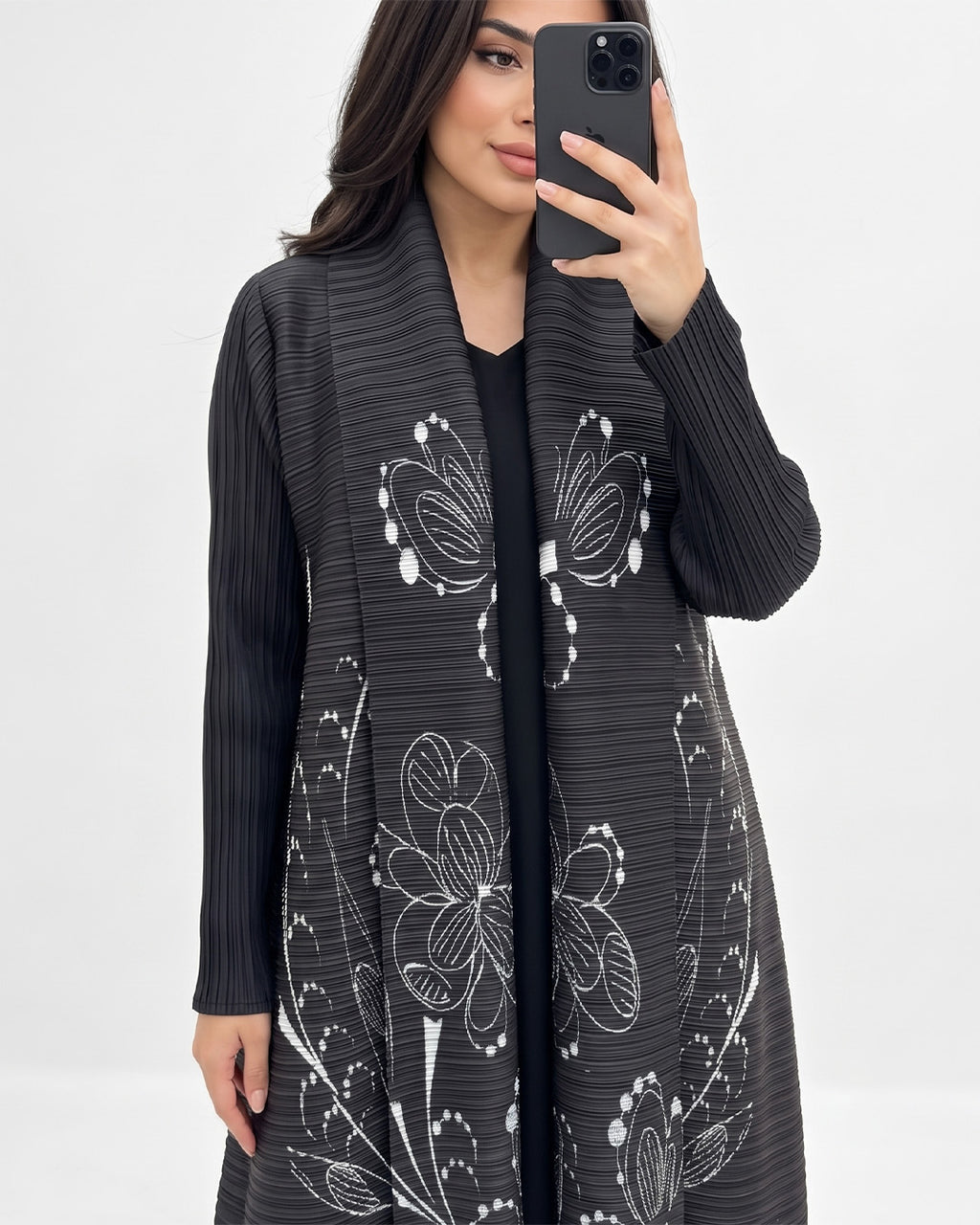 A woman takes a mirror selfie wearing a black pleated open-front cardigan with large white floral line-art patterns, layered over a black top.