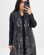 A woman takes a mirror selfie wearing a black pleated open-front cardigan with large white floral line-art patterns, layered over a black top.