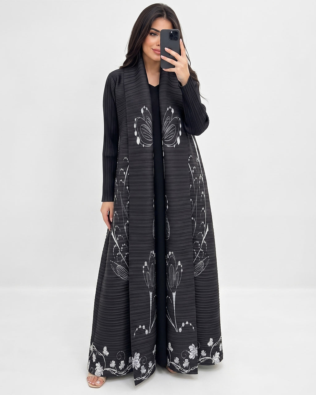 A woman takes a mirror selfie wearing a long black open-front maxi dress with white floral and butterfly patterns. She is standing against a plain white studio background.