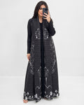 A woman takes a mirror selfie wearing a long black open-front maxi dress with white floral and butterfly patterns. She is standing against a plain white studio background.