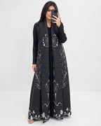 A woman takes a mirror selfie wearing a long black open-front maxi dress with white floral and butterfly patterns. She is standing against a plain white studio background.