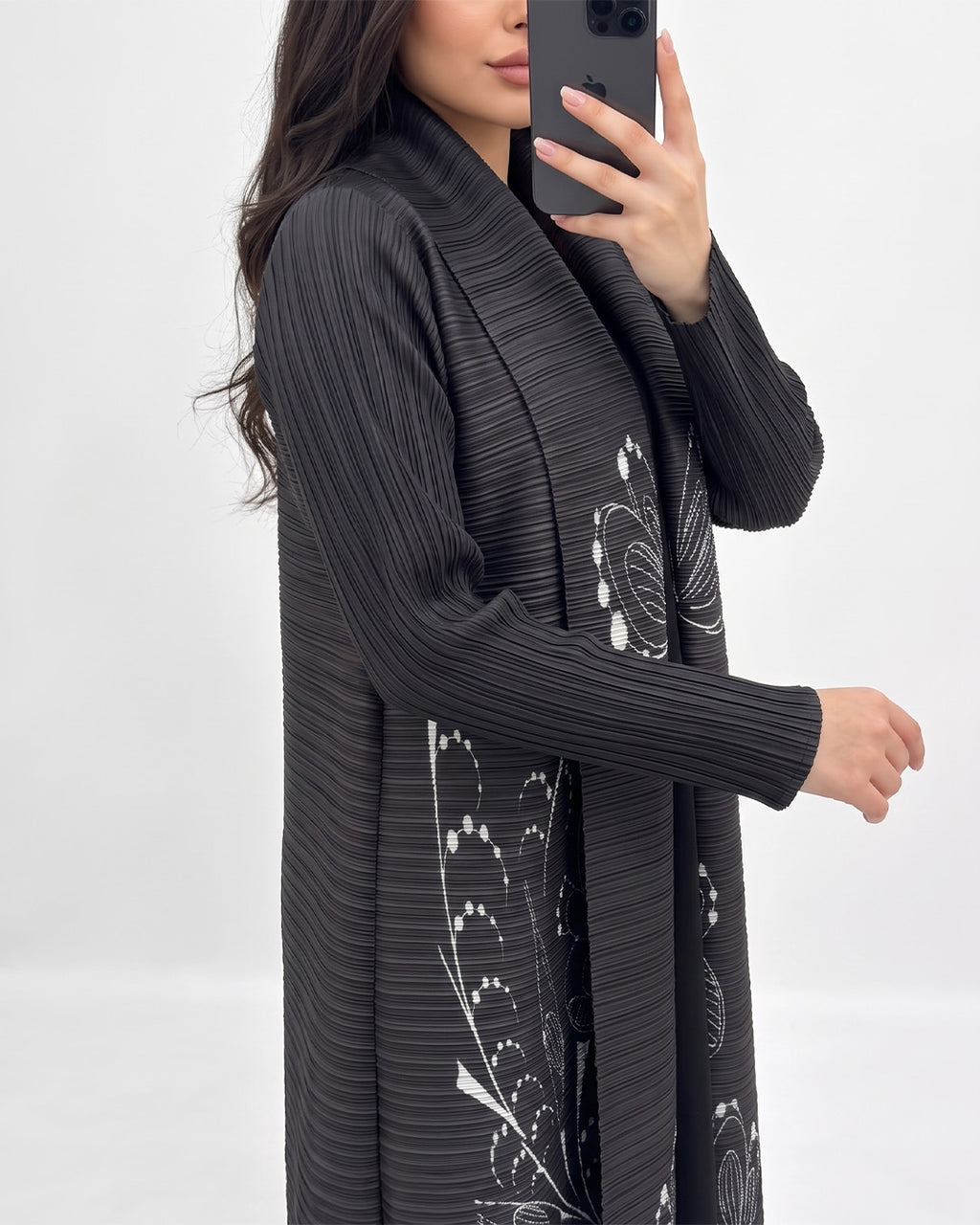 A woman takes a mirror selfie wearing a long, black pleated cardigan with white floral patterns. The background is a clean, neutral white studio setting.