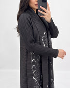 A woman takes a mirror selfie wearing a long, black pleated cardigan with white floral patterns. The background is a clean, neutral white studio setting.