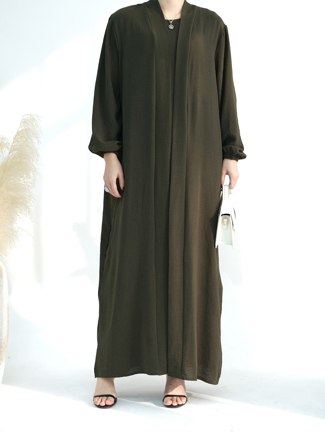 A woman modeling a long, loose-fitting olive green abaya dress with three-quarter sleeves, side pockets, and a V-neckline, paired with black sandals and holding a small white handbag against a bright white studio background.