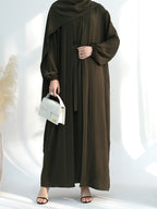 A woman wearing a dark olive brown long-sleeved modest maxi dress with a matching draped headscarf, holding a small white handbag and wearing black flat sandals, set against a bright white background with soft shadows.