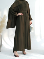 A woman modeling a dark olive green modest maxi dress with a matching draped headscarf, holding a small white clutch and wearing black heeled sandals against a bright white studio background.