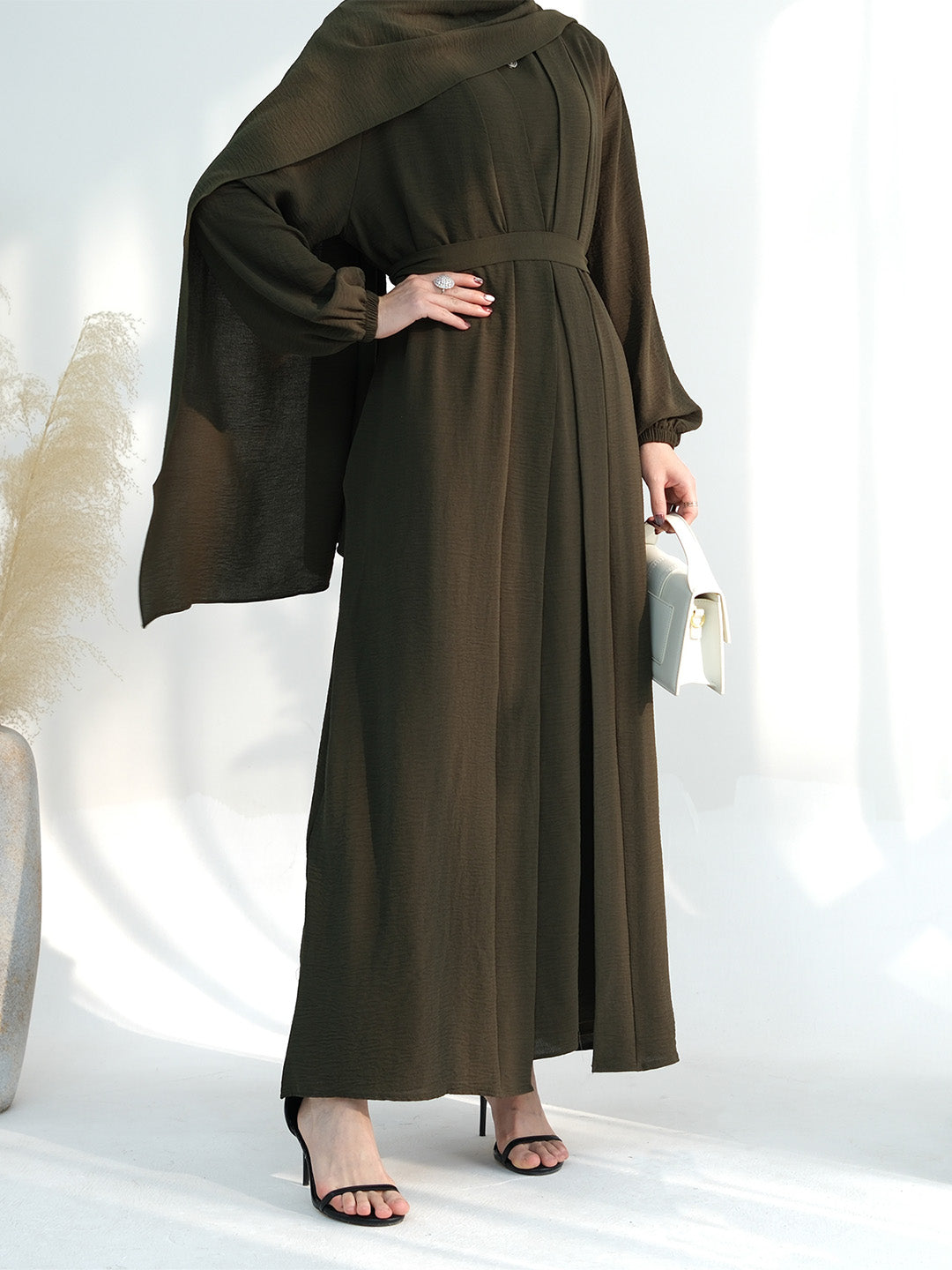 A woman modeling a dark olive green modest maxi dress with a matching draped headscarf, holding a small white clutch and wearing black heeled sandals against a bright white studio background.