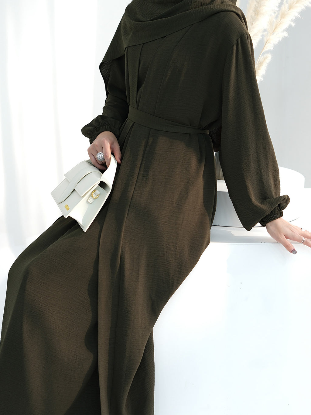 A person wearing a dark olive brown long-sleeve maxi dress with a matching waist belt, holding a white clutch bag, posed against a clean white background.