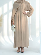 A woman modeling a long beige maxi dress with a draped neckline, long sleeves, and a matching waist tie, styled with a small white handbag and black sandals against a bright white background with soft window shadows.