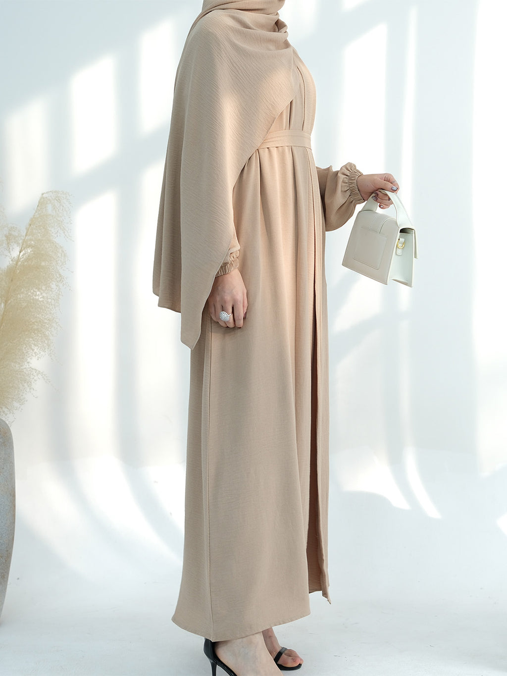 A woman wearing a flowing beige maxi abaya dress with a matching headscarf, holding a small white handbag against a bright white background with soft window shadows.
