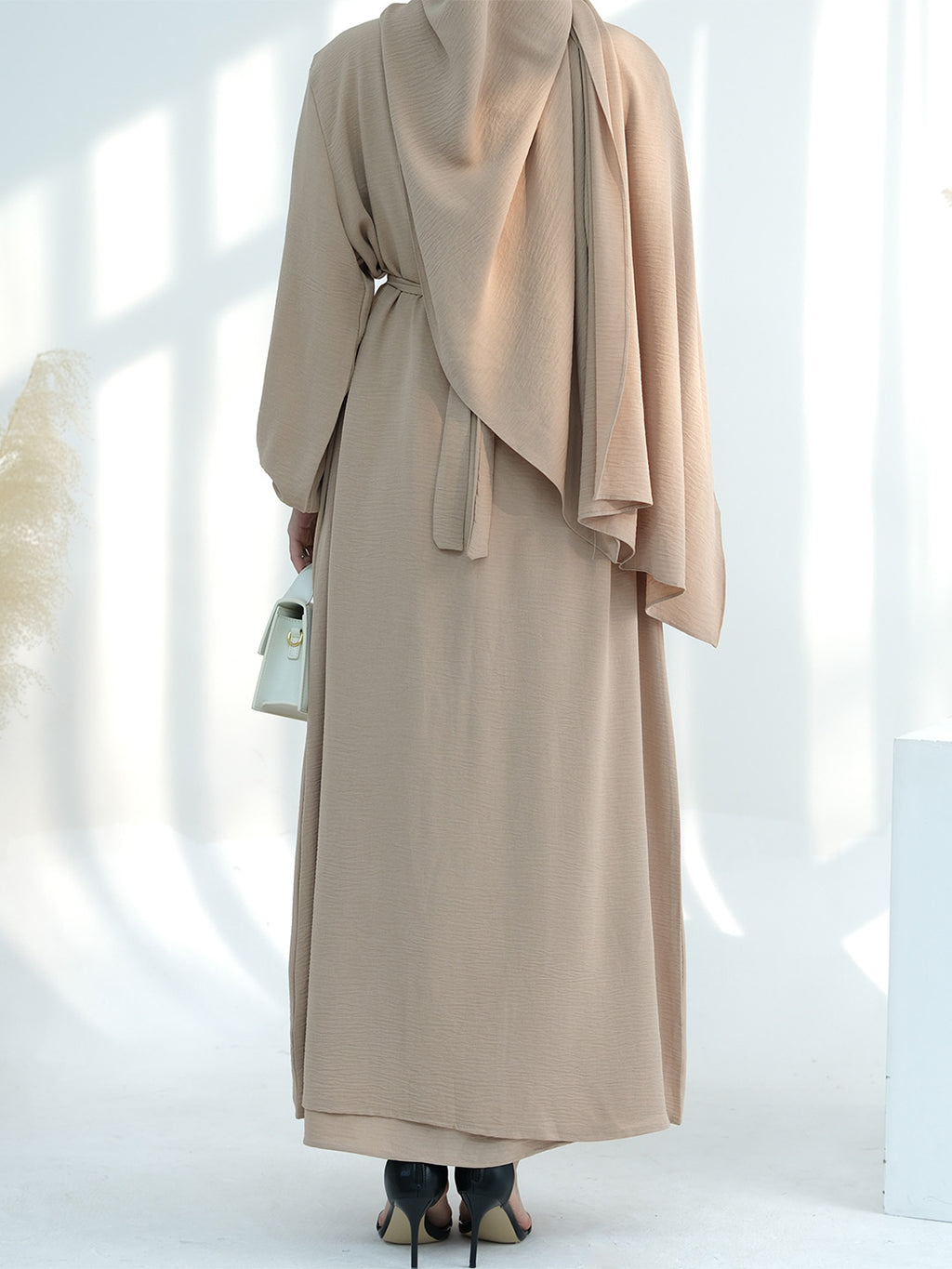 A woman shown from the back wearing a long beige modest dress or abaya with a draped shawl and waist tie, standing in a bright white studio with soft window-like shadows.