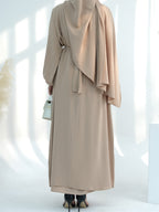A woman shown from the back wearing a long beige modest dress or abaya with a draped shawl and waist tie, standing in a bright white studio with soft window-like shadows.