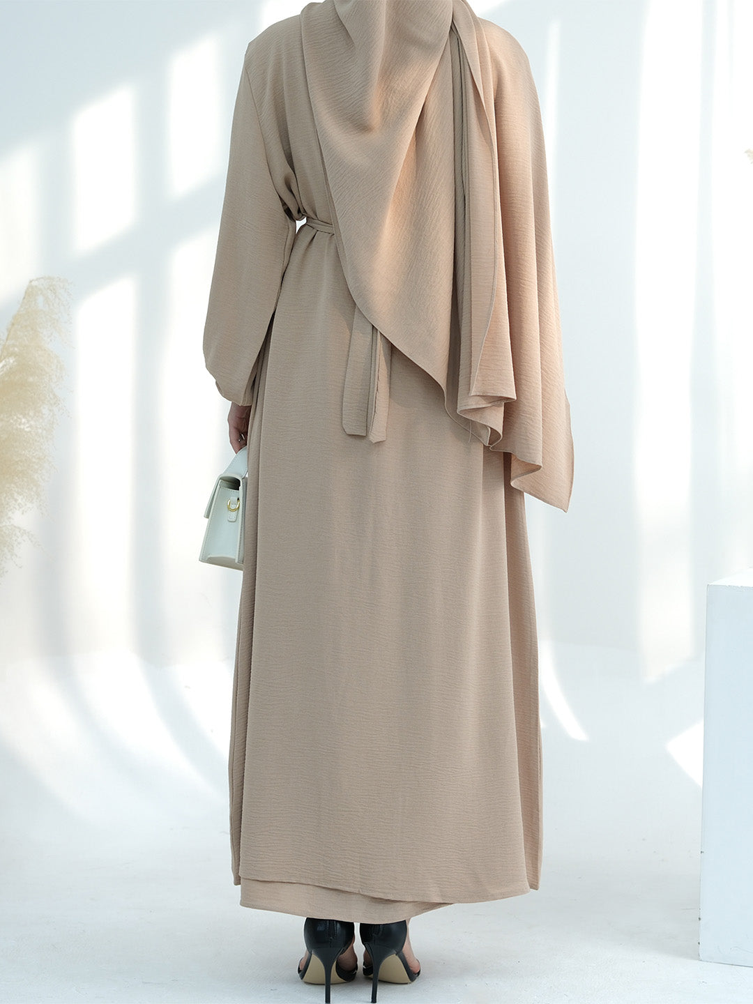 A woman shown from the back wearing a long beige modest dress or abaya with a draped shawl and waist tie, standing in a bright white studio with soft window-like shadows.