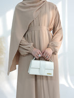 A woman wearing a draped beige abaya dress with a matching belt and a large matching headscarf, holding a structured white handbag with a gold logo plate.