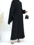 A woman wearing a long black abaya dress with wide draped sleeves and a matching belt, shown from the back while holding a small black handbag.