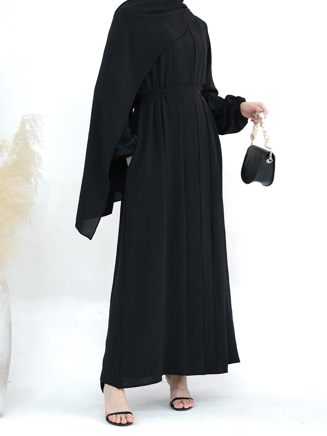 A woman wearing a long black abaya dress with wide draped sleeves and a matching belt, shown from the back while holding a small black handbag.