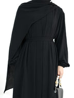 A black long-sleeved abaya dress with a draped shawl and matching waist tie, shown from the back on a model holding a small gray handbag.