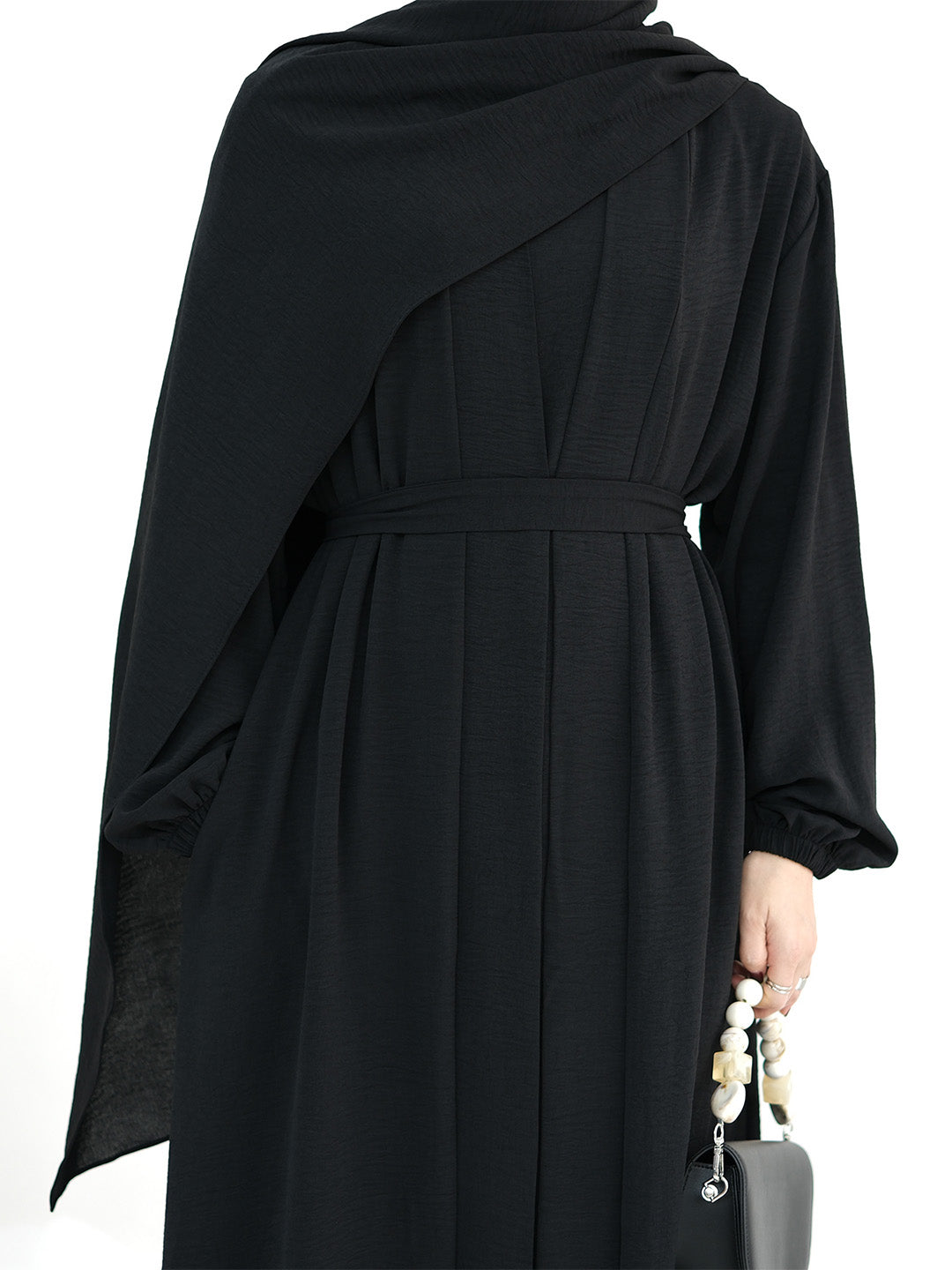 A black long-sleeved abaya dress with a draped shawl and matching waist tie, shown from the back on a model holding a small gray handbag.