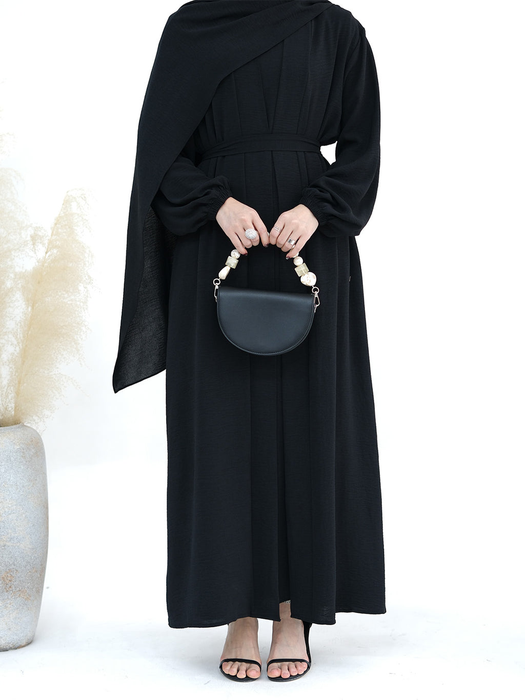 A model wearing a long black draped maxi dress with balloon sleeves, holding a black semi-circular handbag with a pearl handle, set against a white background with a decorative vase of pampas grass on the left.