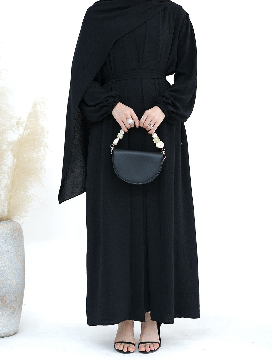 A model wearing a long black draped maxi dress with balloon sleeves, holding a black semi-circular handbag with a pearl handle, set against a white background with a decorative vase of pampas grass on the left.