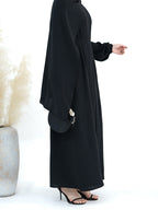 A woman wearing a long black abaya dress with wide draped sleeves and a gathered waist, shown in profile against a clean white background. She is wearing black heeled sandals and holding a small black handbag.