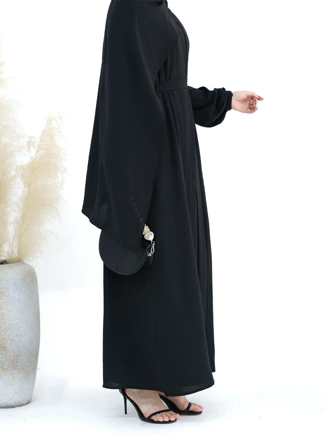 A woman wearing a long black abaya dress with wide draped sleeves and a gathered waist, shown in profile against a clean white background. She is wearing black heeled sandals and holding a small black handbag.