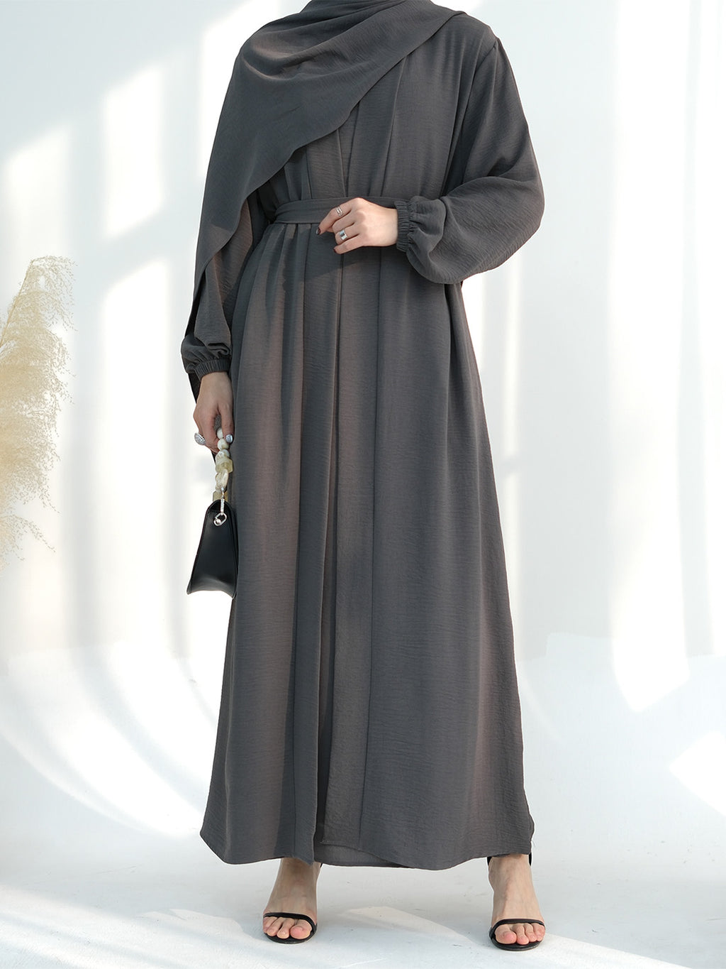 A woman wearing a dark gray maxi abaya dress with a draped shawl, long sleeves, and a matching waist tie, styled with a small black handbag and black sandals.