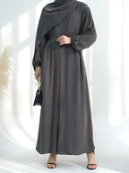 A woman wearing a dark gray maxi abaya dress with a draped shawl, long sleeves, and a matching waist tie, styled with a small black handbag and black sandals.