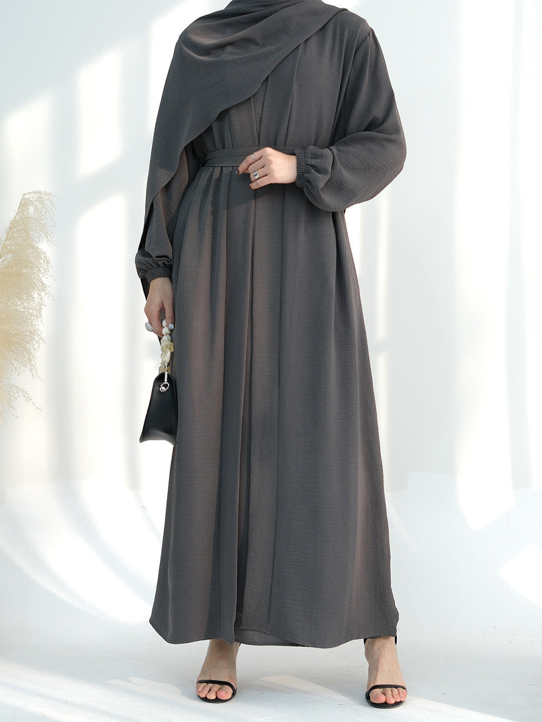 A woman wearing a dark gray maxi abaya dress with a draped shawl, long sleeves, and a matching waist tie, styled with a small black handbag and black sandals.