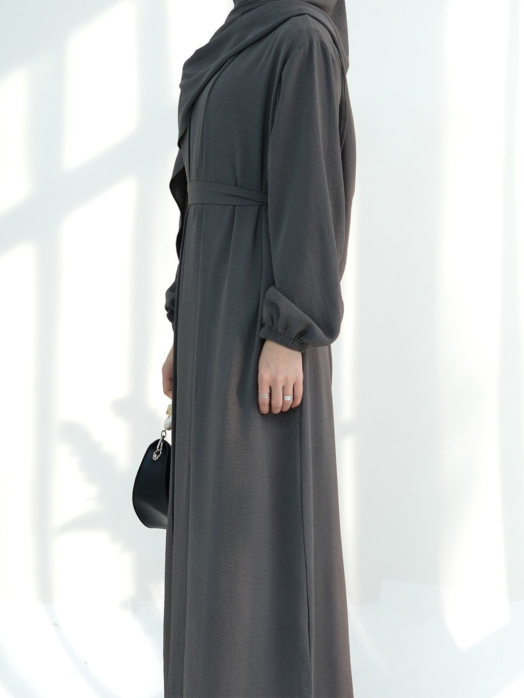 A woman wearing a long dark gray abaya dress with a matching headscarf, shown from the side against a bright white background with soft window shadows.