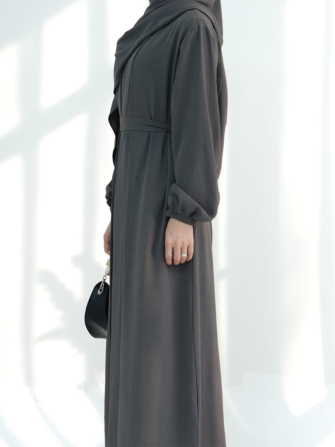 A woman wearing a long dark gray abaya dress with a matching headscarf, shown from the side against a bright white background with soft window shadows.