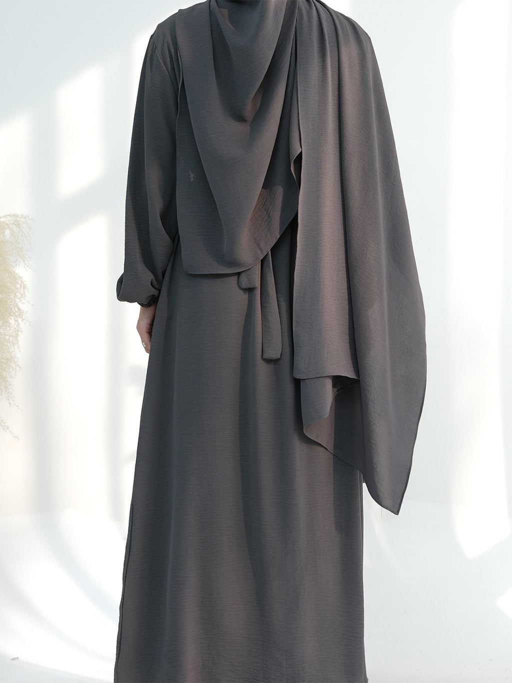 A person wearing a dark charcoal gray abaya dress with a matching draped hijab, shown from the back against a bright white background with soft window shadows.