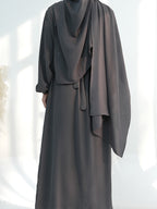 A person wearing a dark charcoal gray abaya dress with a matching draped hijab, shown from the back against a bright white background with soft window shadows.
