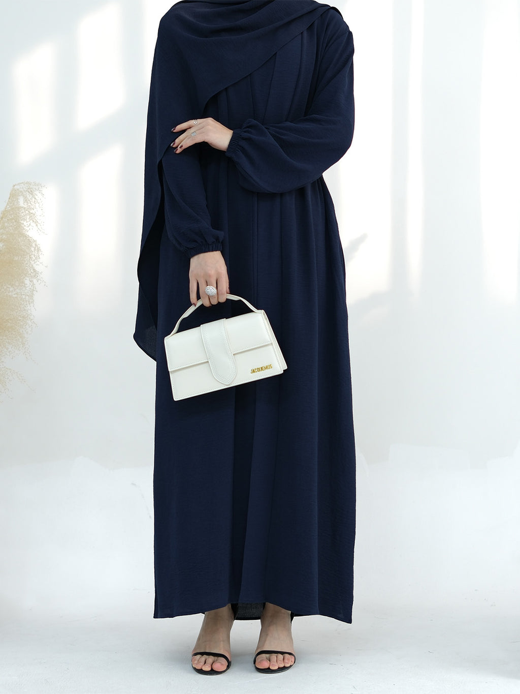 A woman wearing a navy blue long-sleeve maxi abaya dress, holding a structured white handbag, standing against a bright white background with soft window shadows.