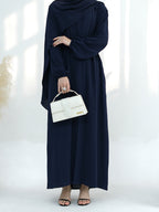 A woman wearing a navy blue long-sleeve maxi abaya dress, holding a structured white handbag, standing against a bright white background with soft window shadows.