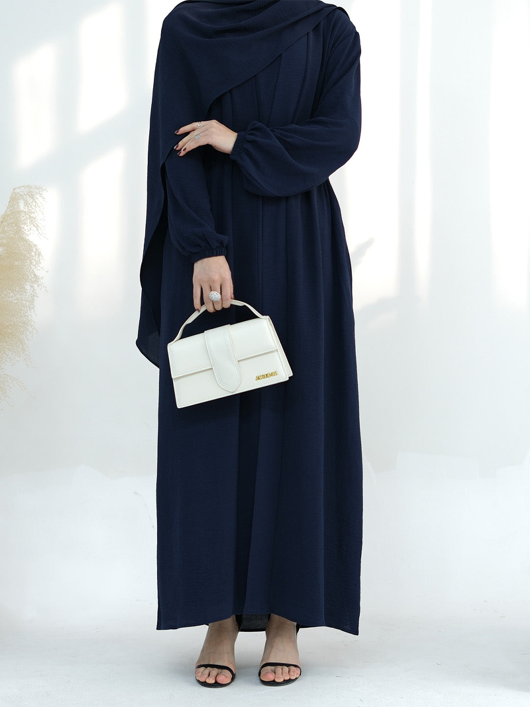 A woman wearing a navy blue long-sleeve maxi abaya dress, holding a structured white handbag, standing against a bright white background with soft window shadows.