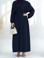 A woman wearing a navy blue long-sleeve maxi dress with an asymmetrical draped neckline and a matching waist tie, holding a small white handbag.