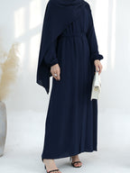 A woman wearing a navy blue maxi abaya dress with a draped shawl, holding a small white handbag and wearing black heeled sandals, set against a bright white background with soft window light shadows.