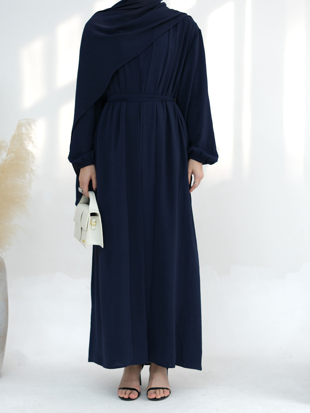 A woman wearing a navy blue long-sleeve maxi dress with an asymmetrical draped neckline and a matching waist tie, holding a small white handbag.
