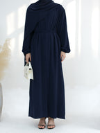 A woman wearing a navy blue long-sleeve maxi dress with an asymmetrical draped neckline and a matching waist tie, holding a small white handbag.