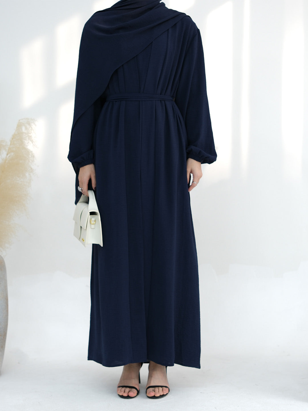 A woman wearing a navy blue long-sleeve maxi dress with an asymmetrical draped neckline and a matching waist tie, holding a small white handbag.