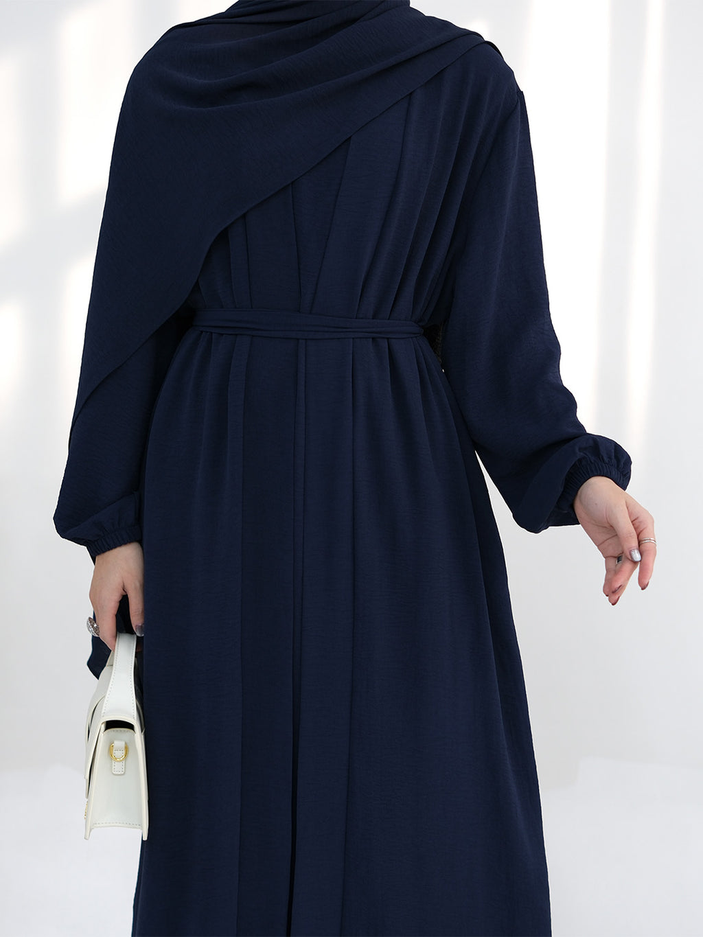 A woman wearing a navy blue long-sleeve maxi dress with a draped asymmetrical shawl collar and a matching waist tie, shown from the back. She is holding a small white handbag against a bright, softly lit background.