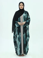 A woman modeling a long black hijab and a matching long abaya with a green and black abstract watercolor pattern, standing against a plain white studio background.