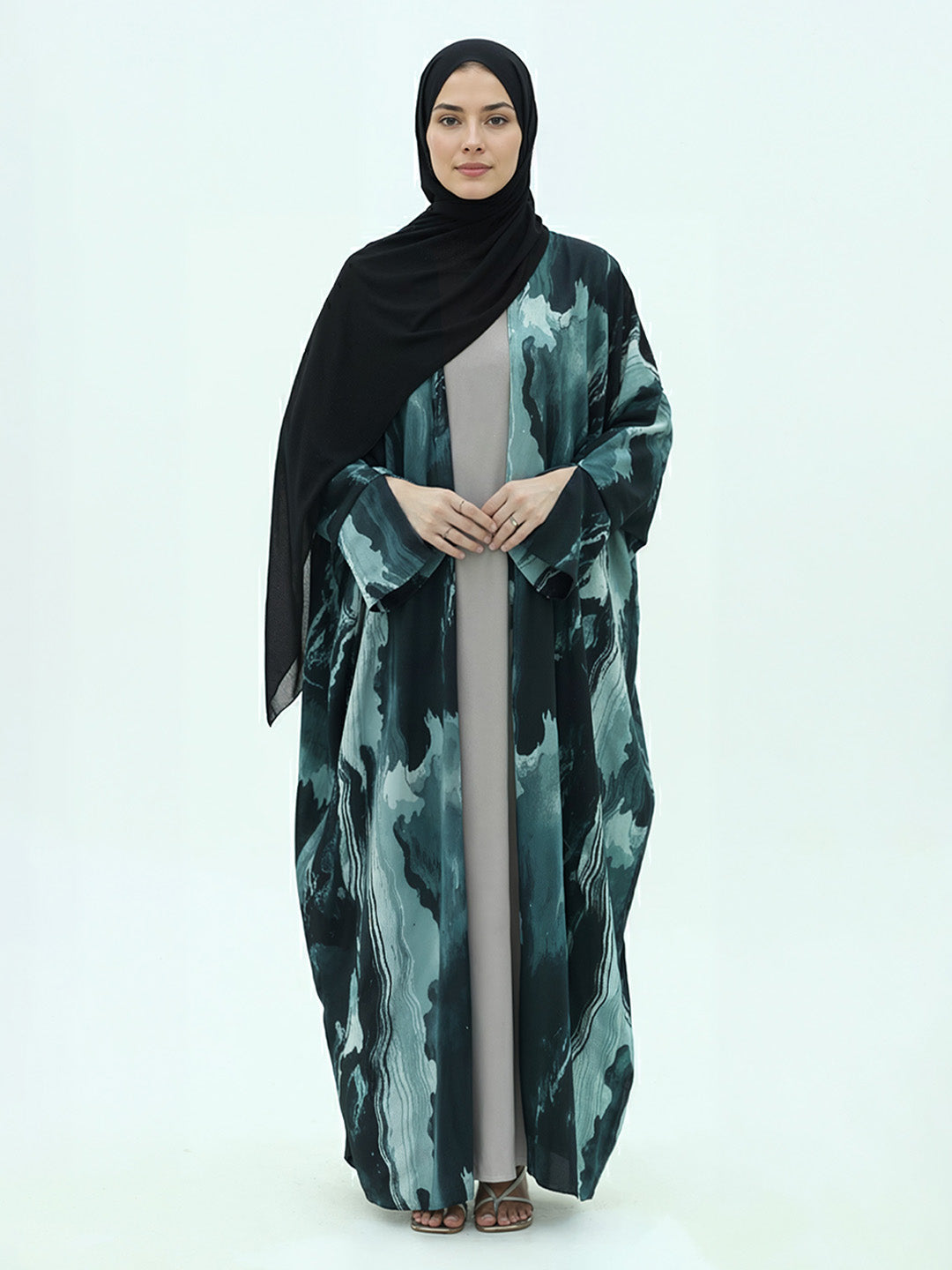 A woman modeling a long black hijab and a matching long abaya with a green and black abstract watercolor pattern, standing against a plain white studio background.