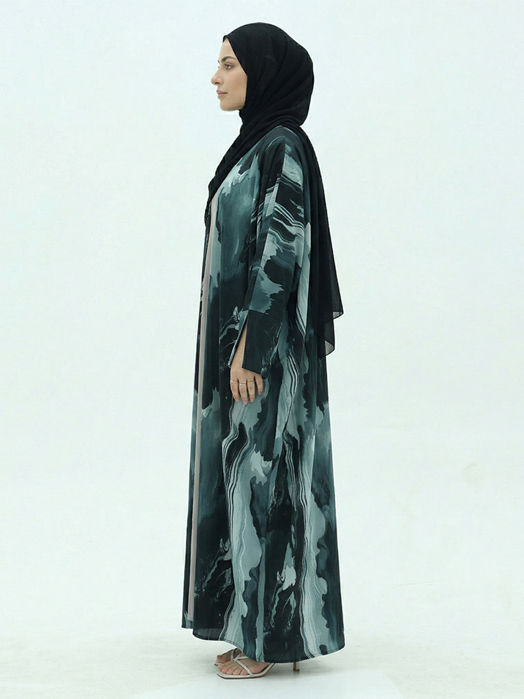 A woman in profile wearing a long black abaya with a green and gray abstract marbled pattern, paired with a solid black hijab, standing against a plain white studio background.