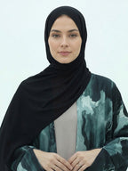 A woman wearing a black hijab and a long-sleeved abaya with a green and black abstract watercolor pattern over a light gray inner top, set against a plain light gray background.